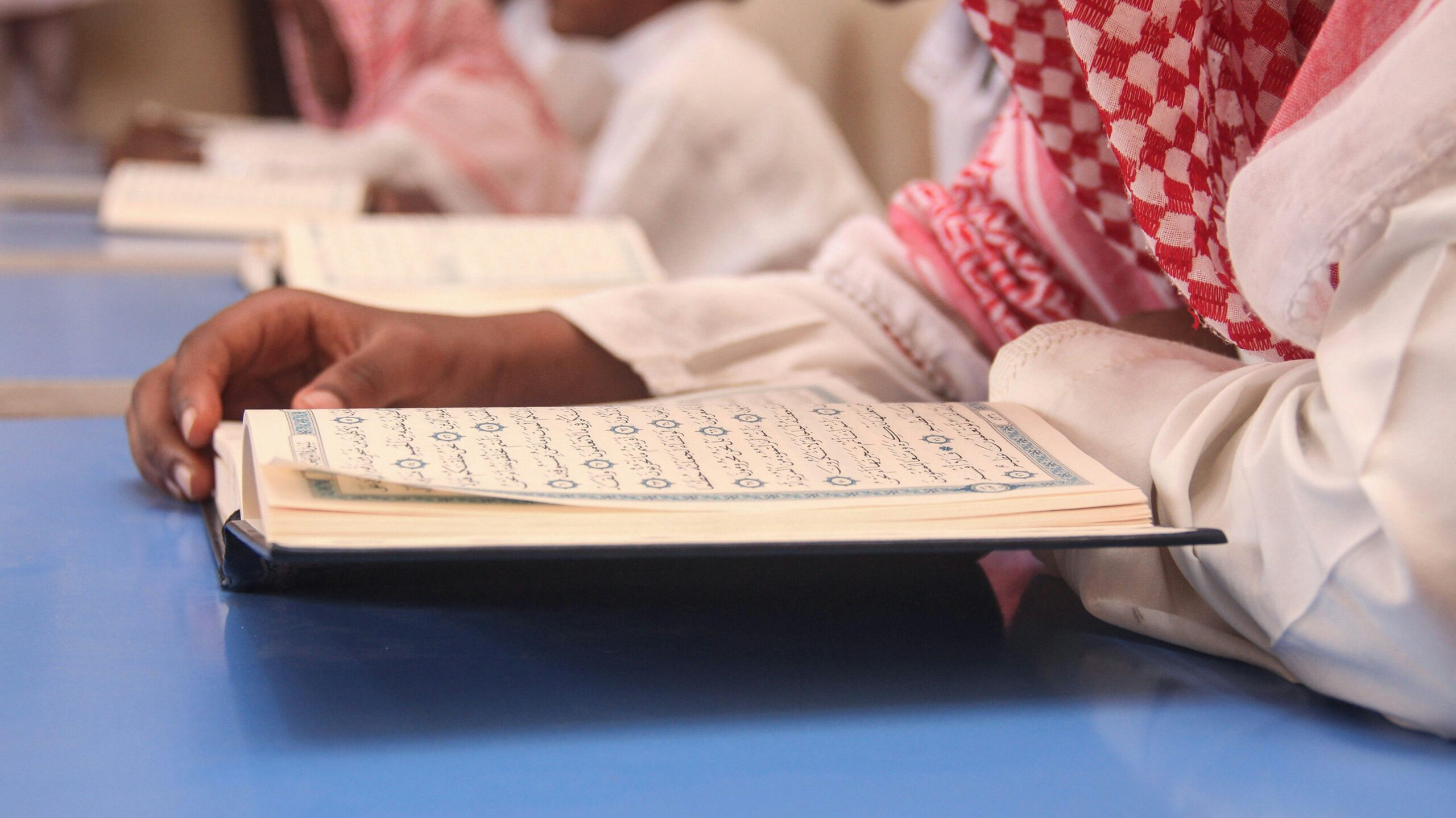 A person in traditional attire studying the Quran indoors, focused on spiritual education.