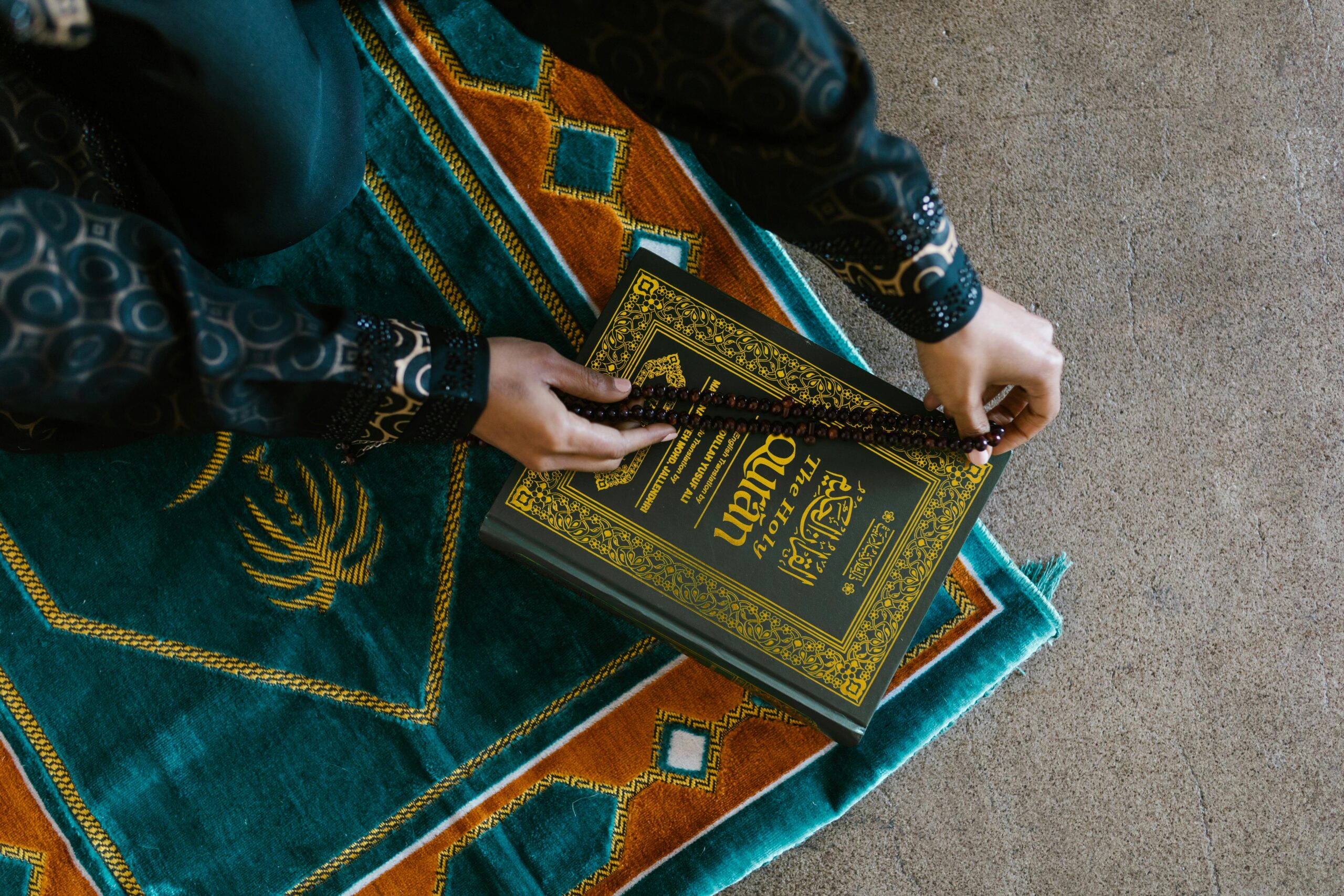 Top view of a person holding prayer beads and Quran on a colorful rug, symbolizing Islamic faith and worship.